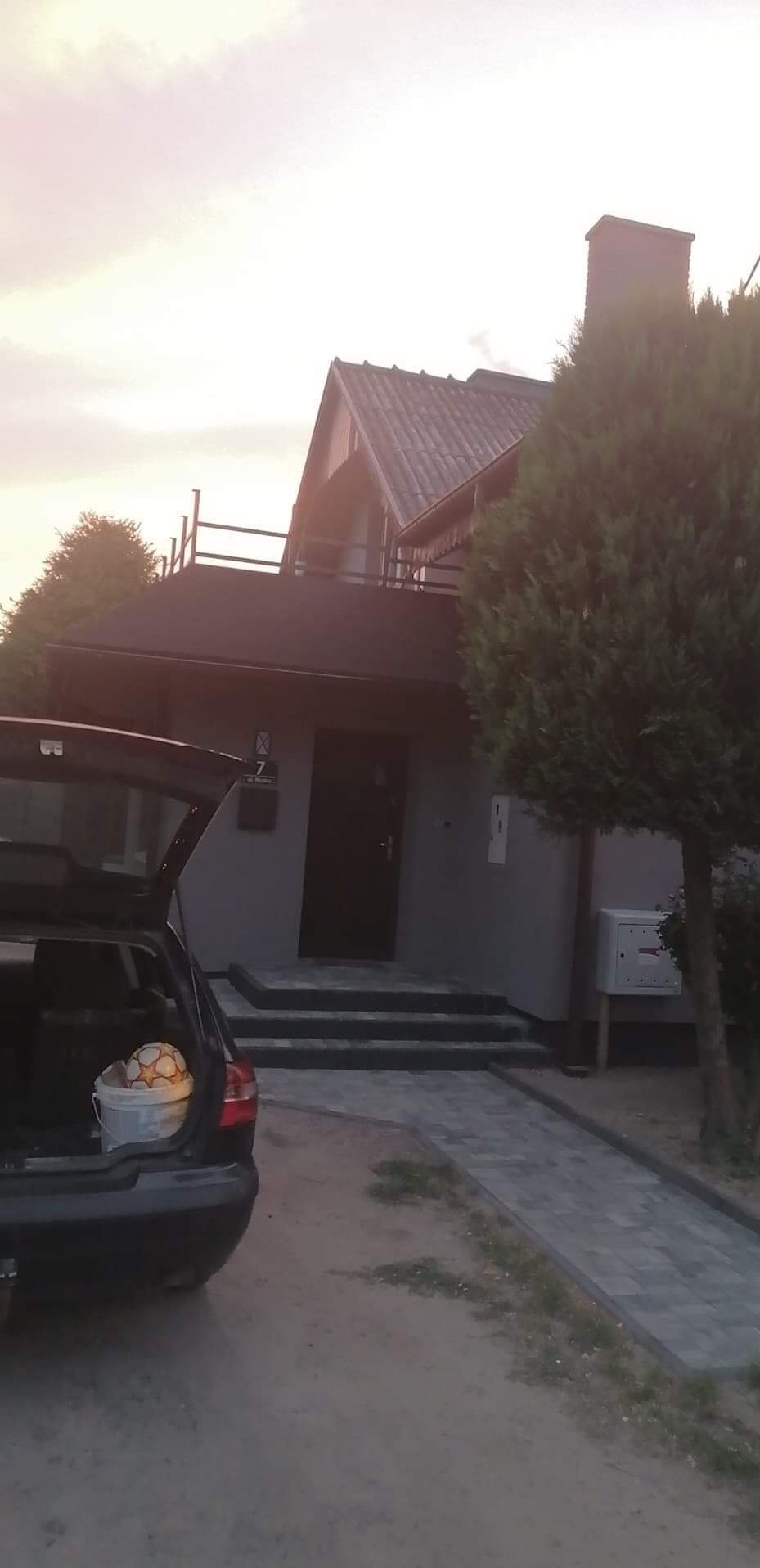 Szary dom jednorodzinny z ciemnym dachem i numerem 7 przy wejściu, widziany z perspektywy otwartego bagażnika samochodu, w którym znajduje się wiadro z piłką.