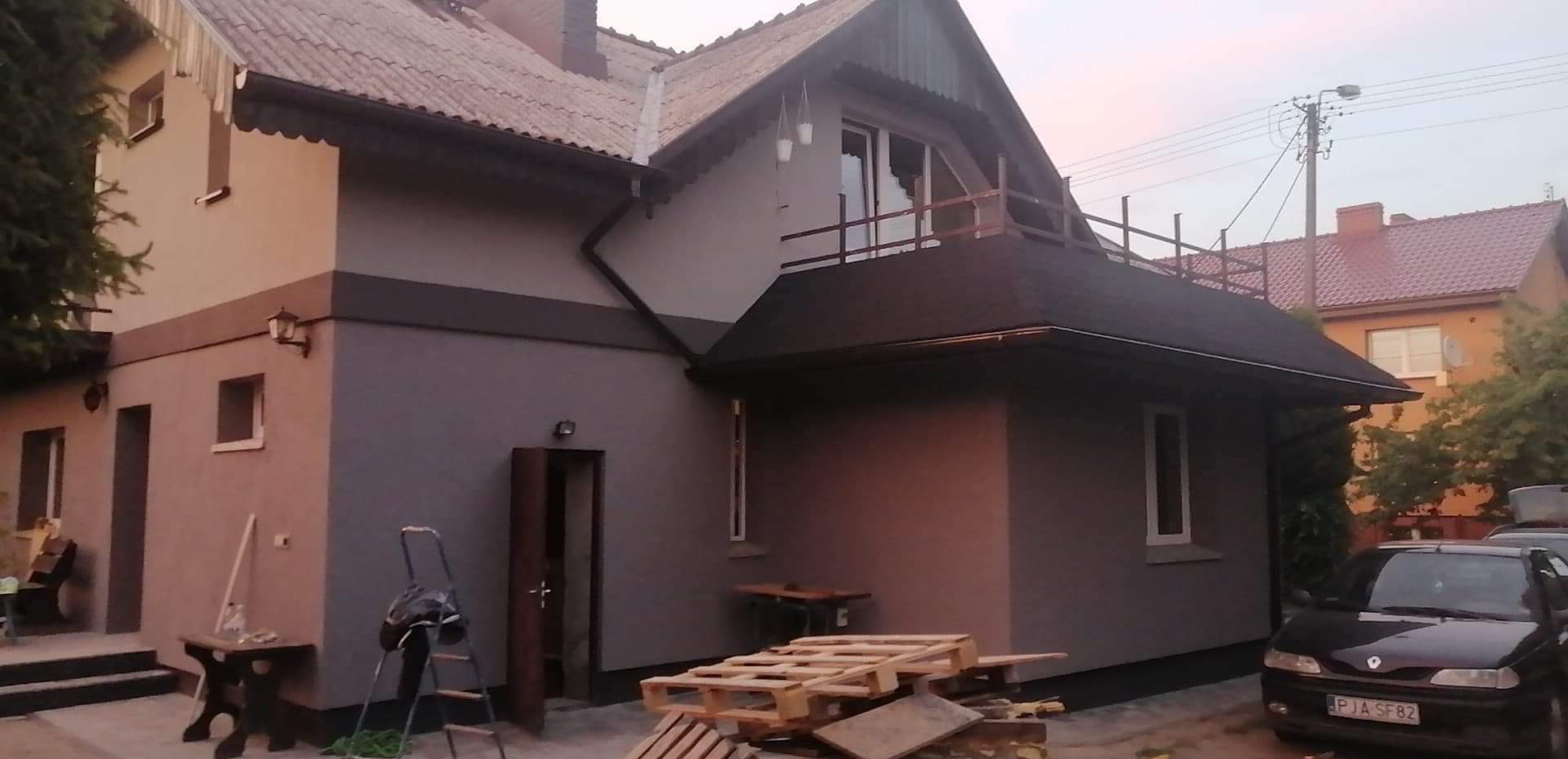 Dwukondygnacyjny dom po remoncie z nową elewacją w odcieniach szarości, widoczne palety i drabina, sugerujące trwające prace wykończeniowe.