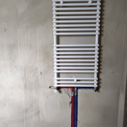 S.G. Instal - Biały grzejnik drabinkowy zamontowany na surowej ścianie, widoczne rury doprowadzające wodę w izolacji niebieskiej i czerwonej, ślady po wierceniu w ścianie.