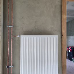 S.G. Instal - Biały, nowy grzejnik panelowy zamontowany na surowej, szarej ścianie, z widocznymi miedzianymi rurami i uchwytami, w tle fragment skrzynki narzędziowej.