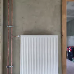Biały, nowy grzejnik panelowy zamontowany na surowej, szarej ścianie, z widocznymi miedzianymi rurami i uchwytami, w tle fragment skrzynki narzędziowej.
