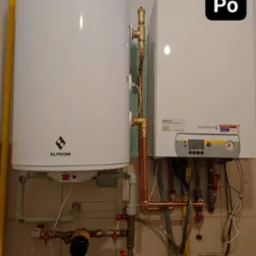 Biały bojler marki Elprom i wiszący piec gazowy De Dietrich z miedzianą instalacją hydrauliczną na tle beżowych płytek.