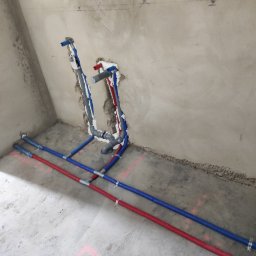 S.G. Instal - Surowe wnętrze łazienki w trakcie instalacji wodno-kanalizacyjnej, widoczne pionowe i poziome rury w kolorach niebieskim i czerwonym, zabezpieczone pianką montażową przy ścianie, różowe oznaczenia...