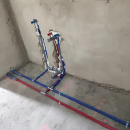 Surowe wnętrze łazienki w trakcie instalacji wodno-kanalizacyjnej, widoczne pionowe i poziome rury w kolorach niebieskim i czerwonym, zabezpieczone pianką montażową przy ścianie, różowe oznaczenia...