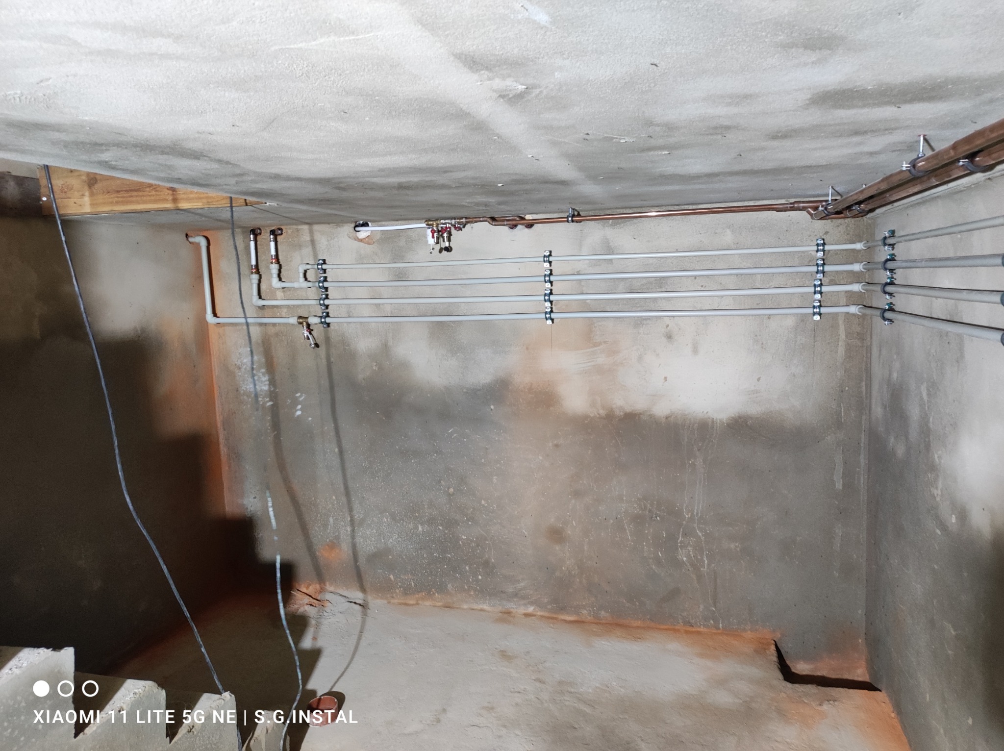 Szare rury instalacji wodnej i miedziane rury grzewcze zamocowane do sufitu w surowym pomieszczeniu z betonowymi ścianami i podłogą. Widoczne elementy mocujące rury.