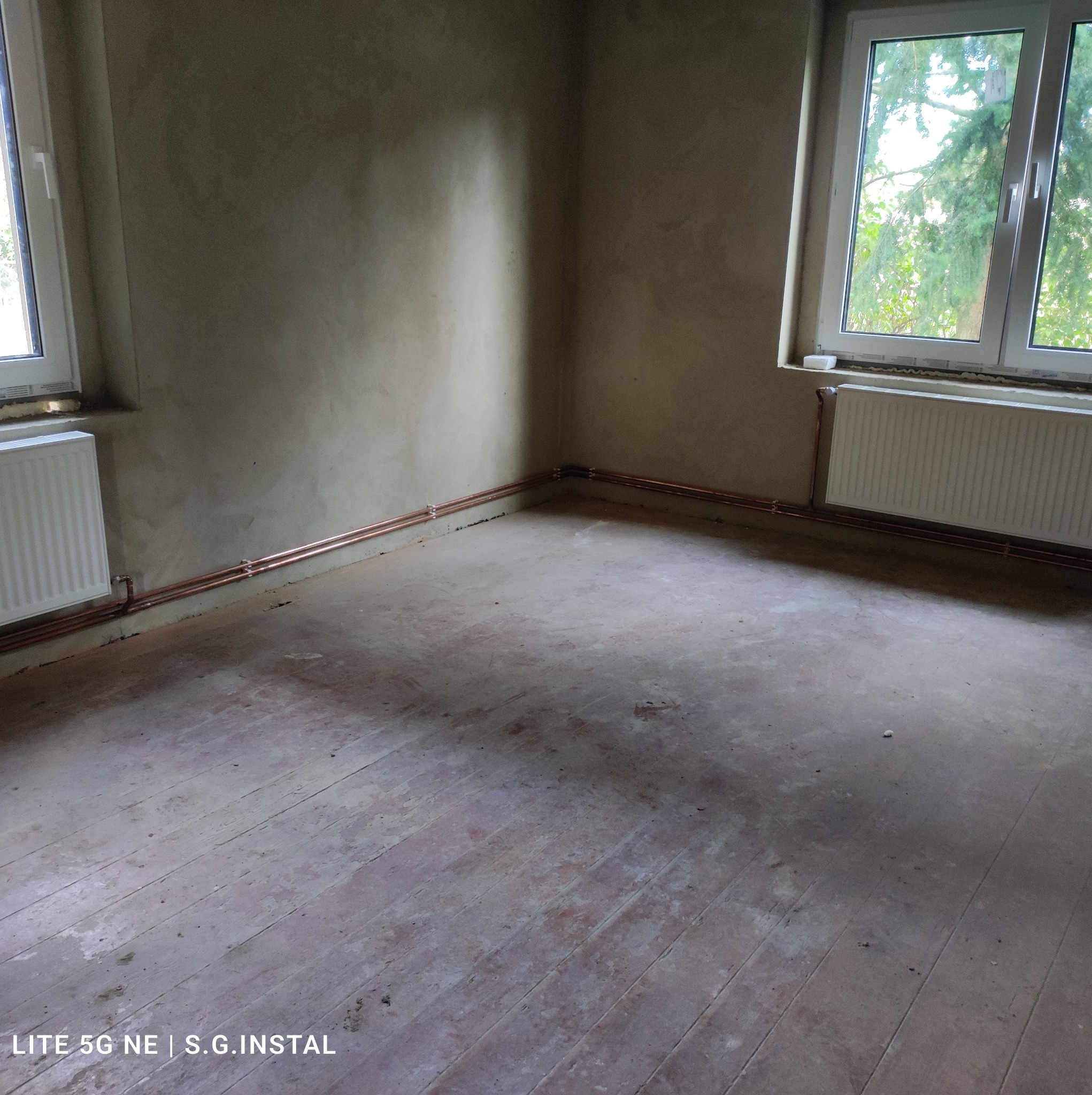 Surowe wnętrze pokoju z nową instalacją centralnego ogrzewania: miedziane rury grzewcze biegnące wzdłuż ścian pod oknami, grzejniki panelowe zamontowane pod oknami, gołe ściany przygotowane...