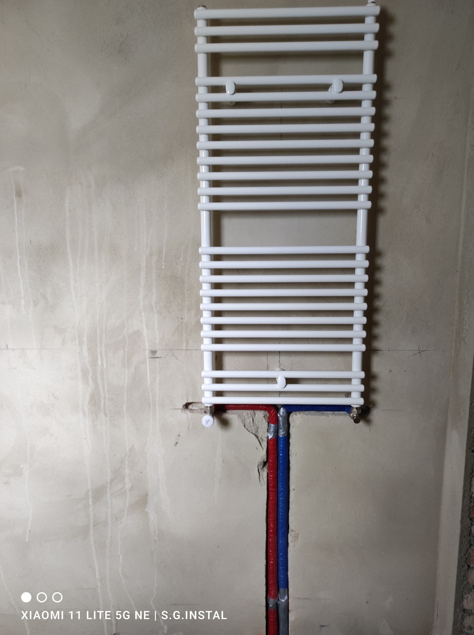 Biały grzejnik drabinkowy zamontowany na surowej ścianie, widoczne rury doprowadzające wodę w izolacji niebieskiej i czerwonej, ślady po wierceniu w ścianie.