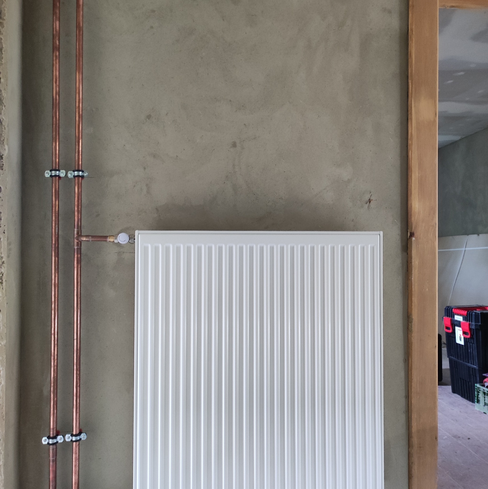 Biały, nowy grzejnik panelowy zamontowany na surowej, szarej ścianie, z widocznymi miedzianymi rurami i uchwytami, w tle fragment skrzynki narzędziowej.