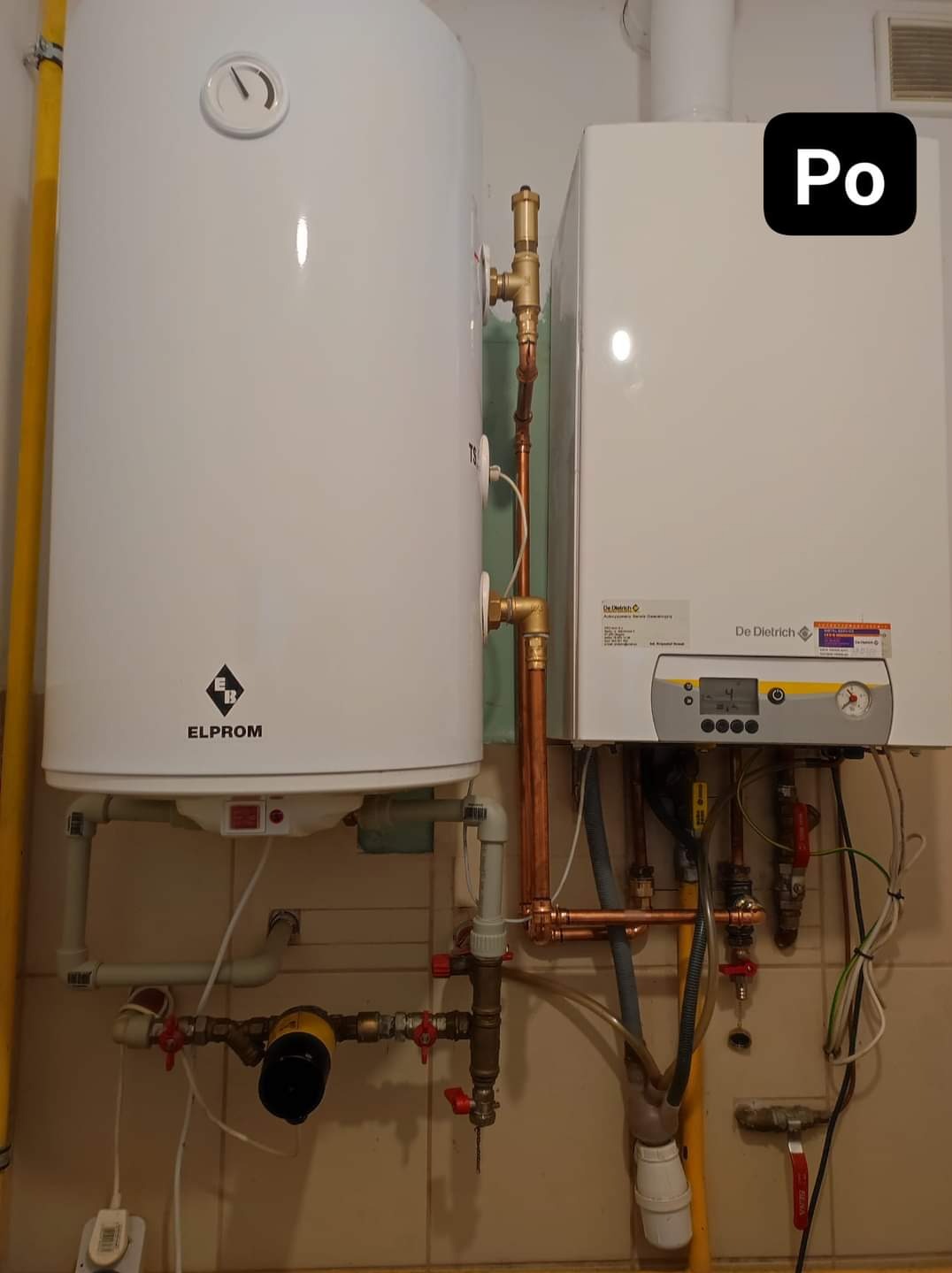 Biały bojler marki Elprom i wiszący piec gazowy De Dietrich z miedzianą instalacją hydrauliczną na tle beżowych płytek.