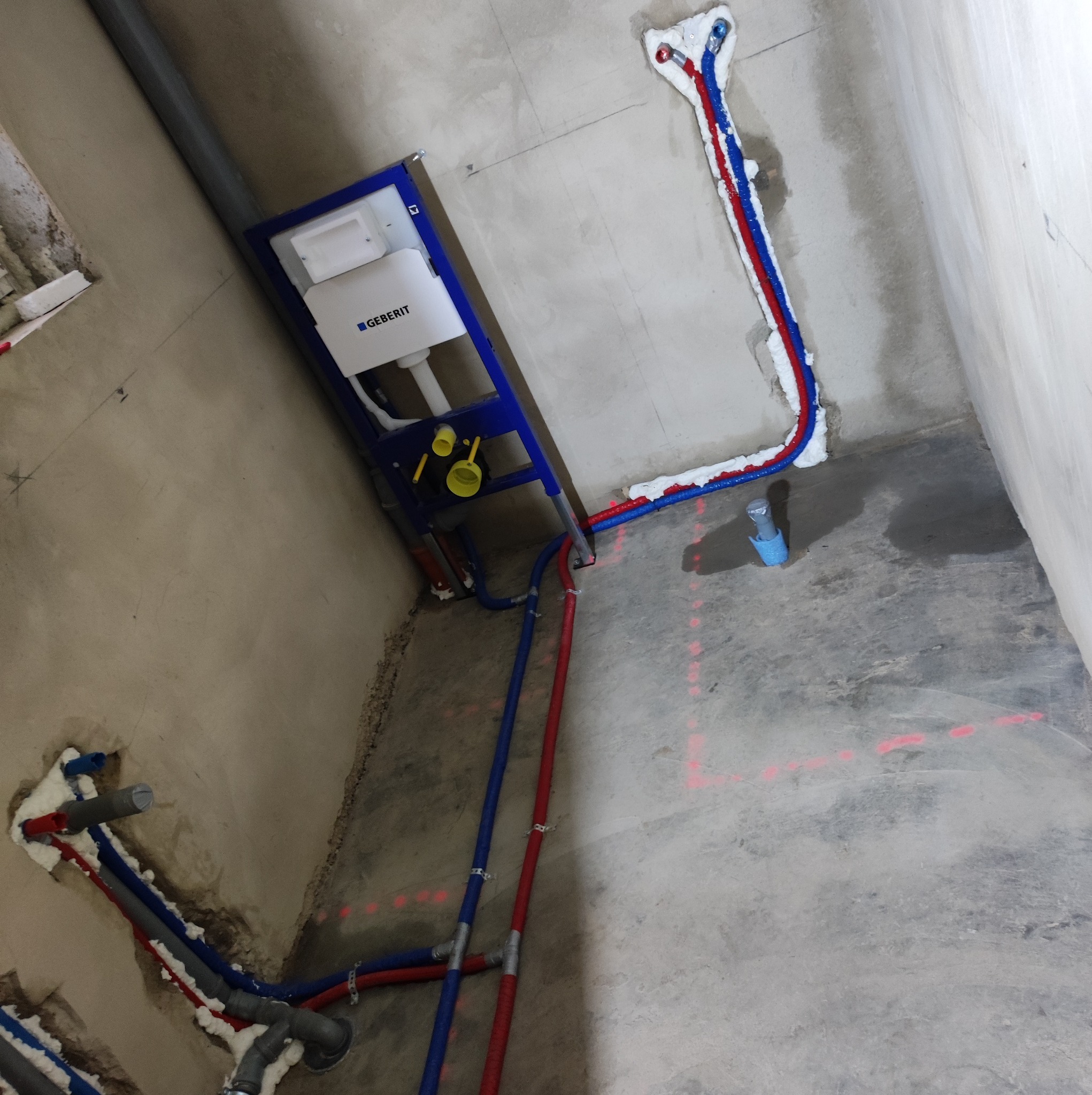 Montaż podtynkowego stelaża WC Geberit z widocznymi przyłączami wodnymi w kolorze czerwonym i niebieskim, zabezpieczonymi pianką montażową w surowym stanie wykończenia łazienki.