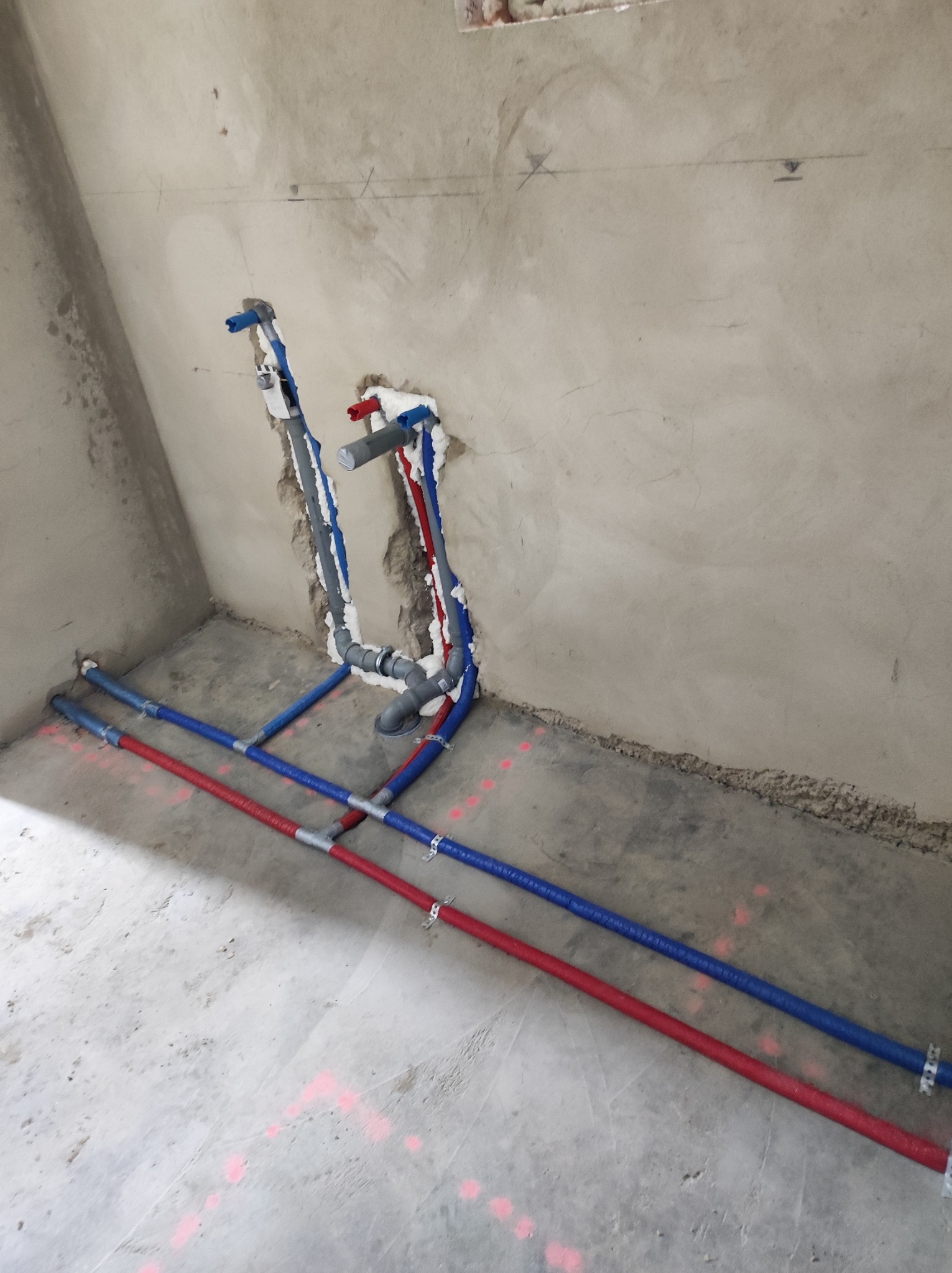 Surowe wnętrze łazienki w trakcie instalacji wodno-kanalizacyjnej, widoczne pionowe i poziome rury w kolorach niebieskim i czerwonym, zabezpieczone pianką montażową przy ścianie, różowe oznaczenia...