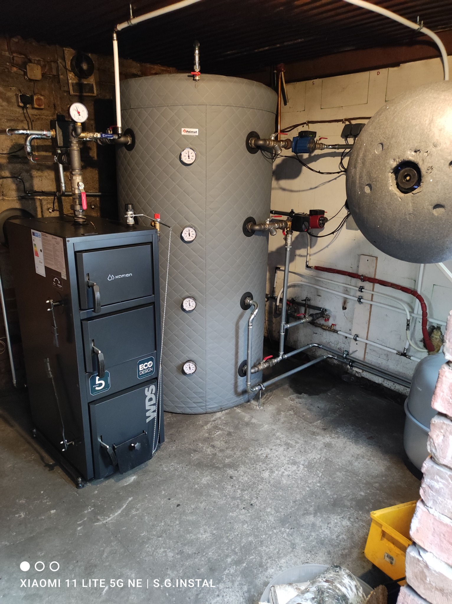 Kotłownia z piecem Kamen Eco Design WDS, zbiornikiem akumulacyjnym Polmet i instalacją grzewczą w piwnicy z ceglaną ścianą w tle.