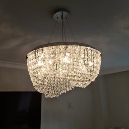 Elektro-Instal Krzysztof Muszyński - Kryształowy żyrandol z metalową obręczą zawieszony na cienkich linkach, oświetlający wnętrze, widoczne ślady po przewodach na ścianie.