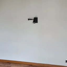 Elektro-Instal Krzysztof Muszyński - Czarna, regulowana lampa ścienna z dwoma reflektorami zamontowana na białej ścianie, widoczna listwa przypodłogowa i gniazdko elektryczne w dolnym rogu.