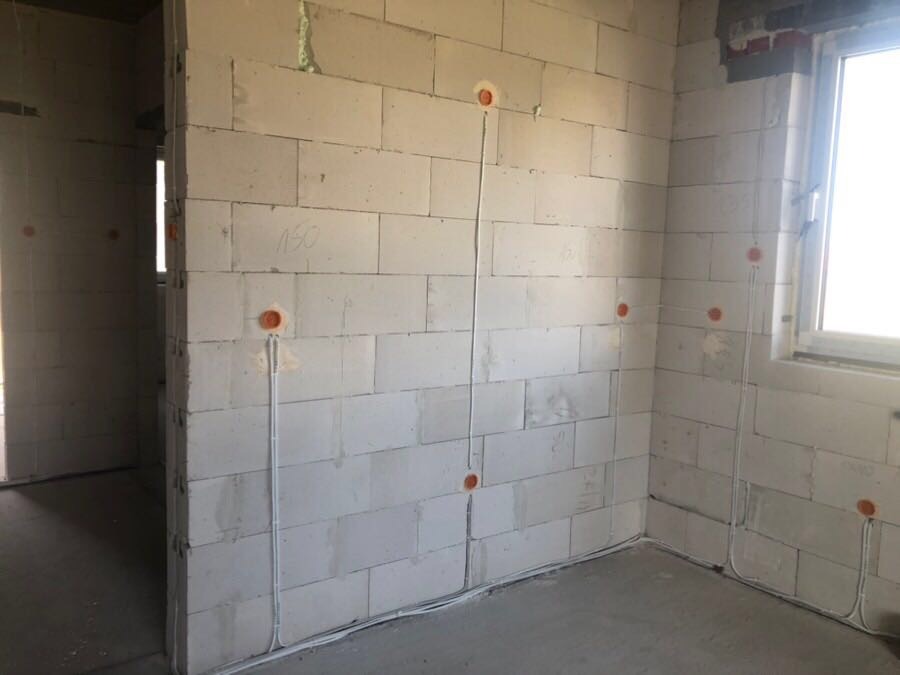 Wnętrze w stanie surowym z widocznymi przewodami elektrycznymi poprowadzonymi wzdłuż ścian z bloczków betonowych do puszek instalacyjnych.