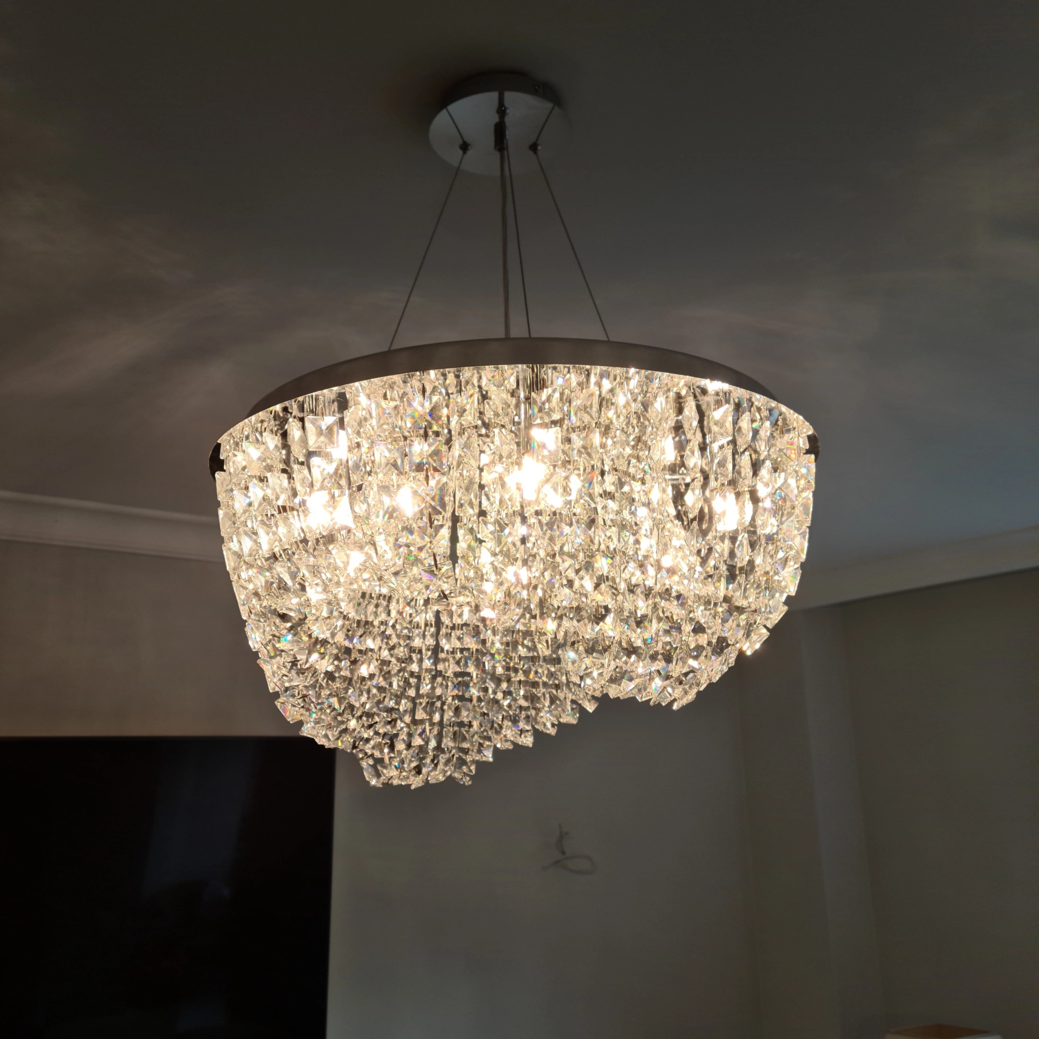 Kryształowy żyrandol z metalową obręczą zawieszony na cienkich linkach, oświetlający wnętrze, widoczne ślady po przewodach na ścianie.