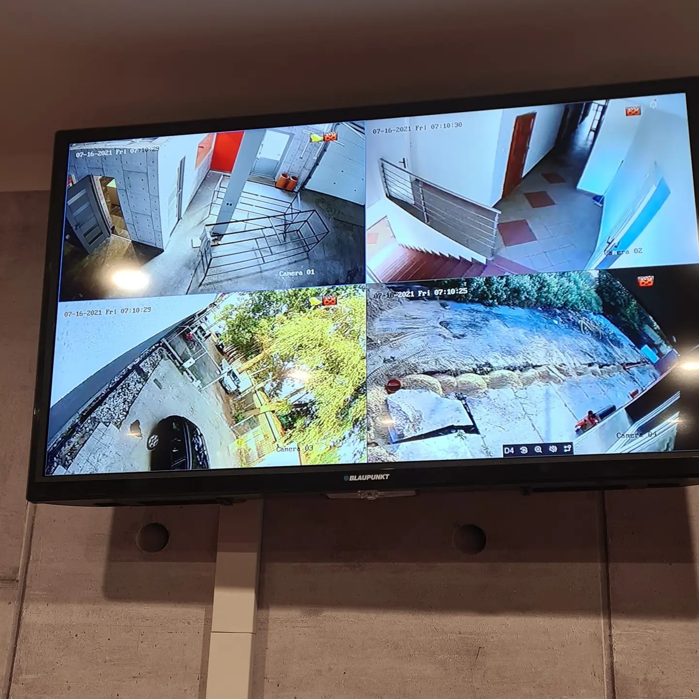 Monitor wyświetlający obraz z czterech kamer monitoringu: widok na klatkę schodową, podwórko i wejście do budynku.