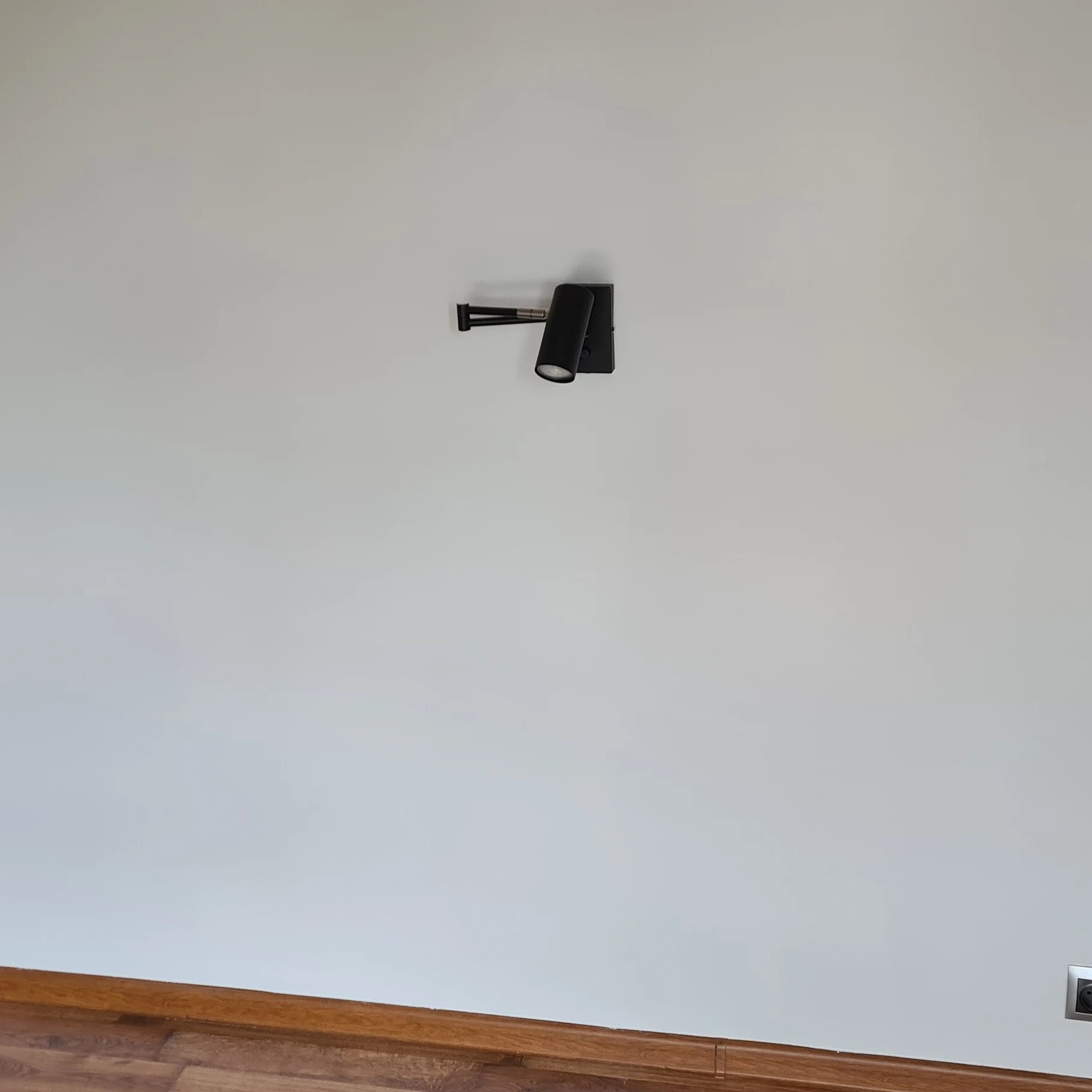 Czarna, regulowana lampa ścienna z dwoma reflektorami zamontowana na białej ścianie, widoczna listwa przypodłogowa i gniazdko elektryczne w dolnym rogu.