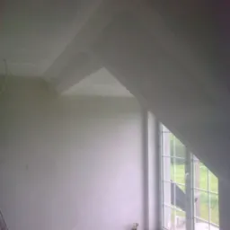 Poddasze w trakcie remontu, ściany i sufit po szpachlowaniu, widoczne okno dachowe, drabina oparta o ścianę, mała figurka przy gniazdku.