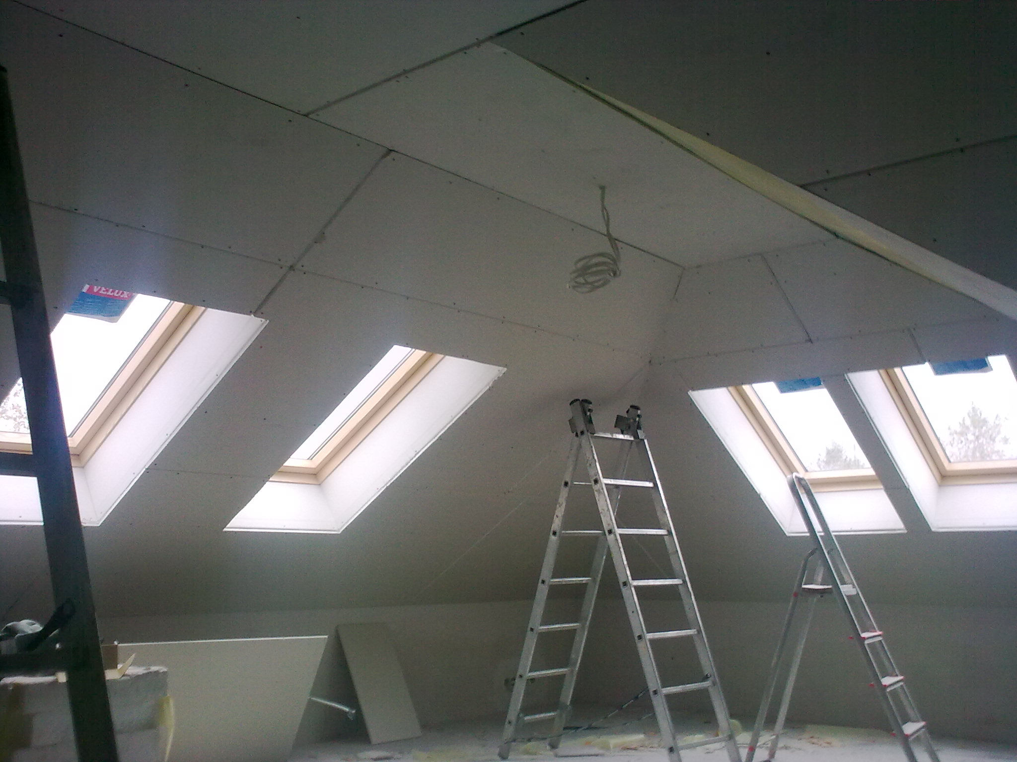 Poddasze z oknami dachowymi, pokryte płytami kartonowo-gipsowymi, widoczne dwie aluminiowe drabiny i zwisający przewód elektryczny.