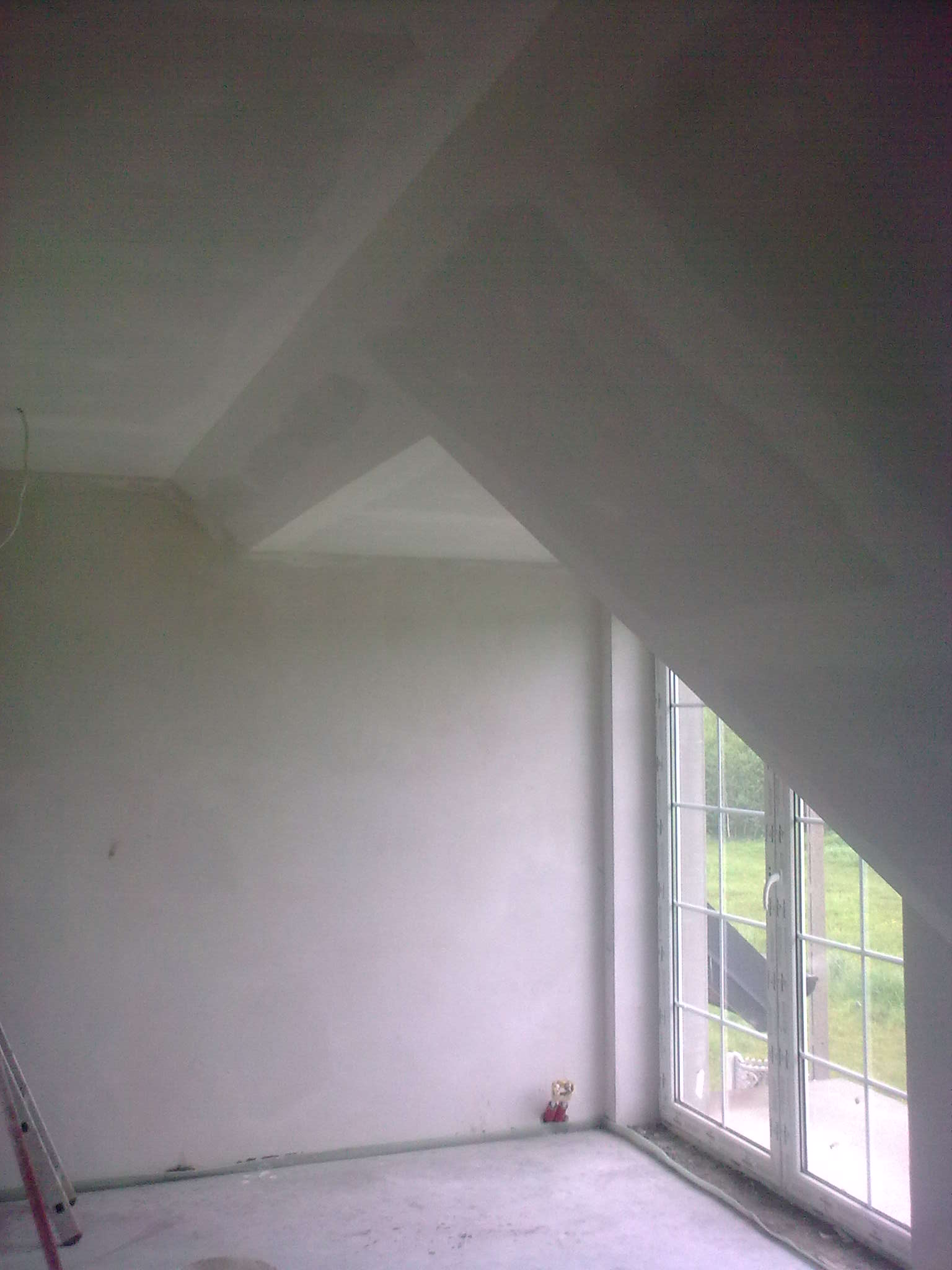 Poddasze w trakcie remontu, ściany i sufit po szpachlowaniu, widoczne okno dachowe, drabina oparta o ścianę, mała figurka przy gniazdku.