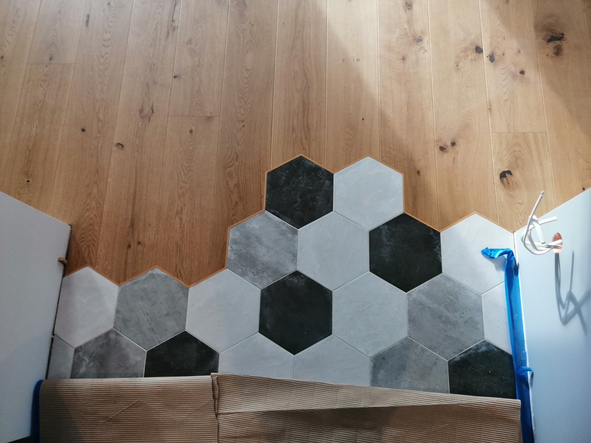 Połączenie drewnianej podłogi z szarą, heksagonalną mozaiką podłogową, zabezpieczone folią malarską przy ścianie.