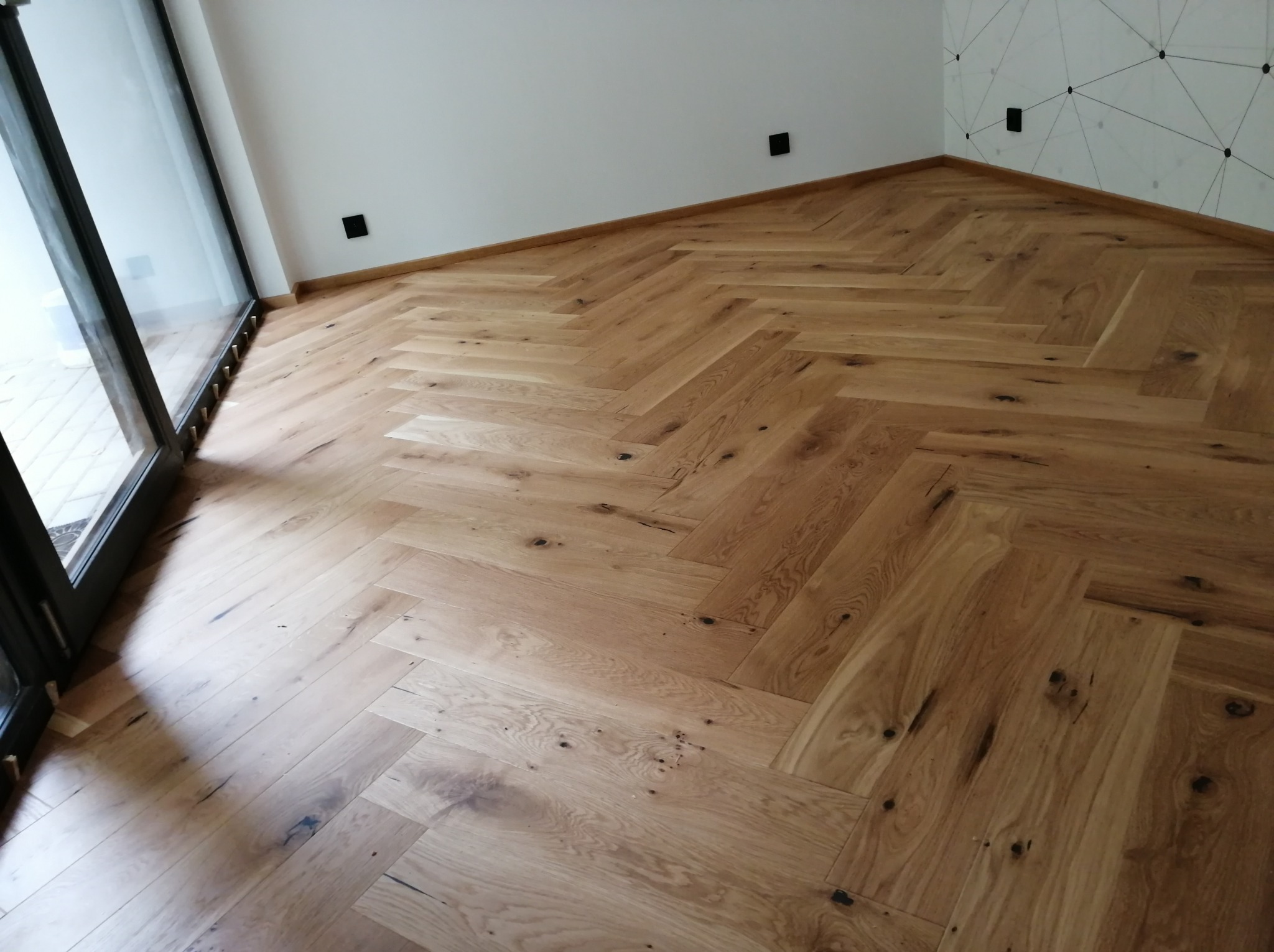 Jasny parkiet ułożony we wzór jodełki francuskiej w nowym pokoju z czarnymi gniazdkami elektrycznymi i minimalistyczną dekoracją ściany.