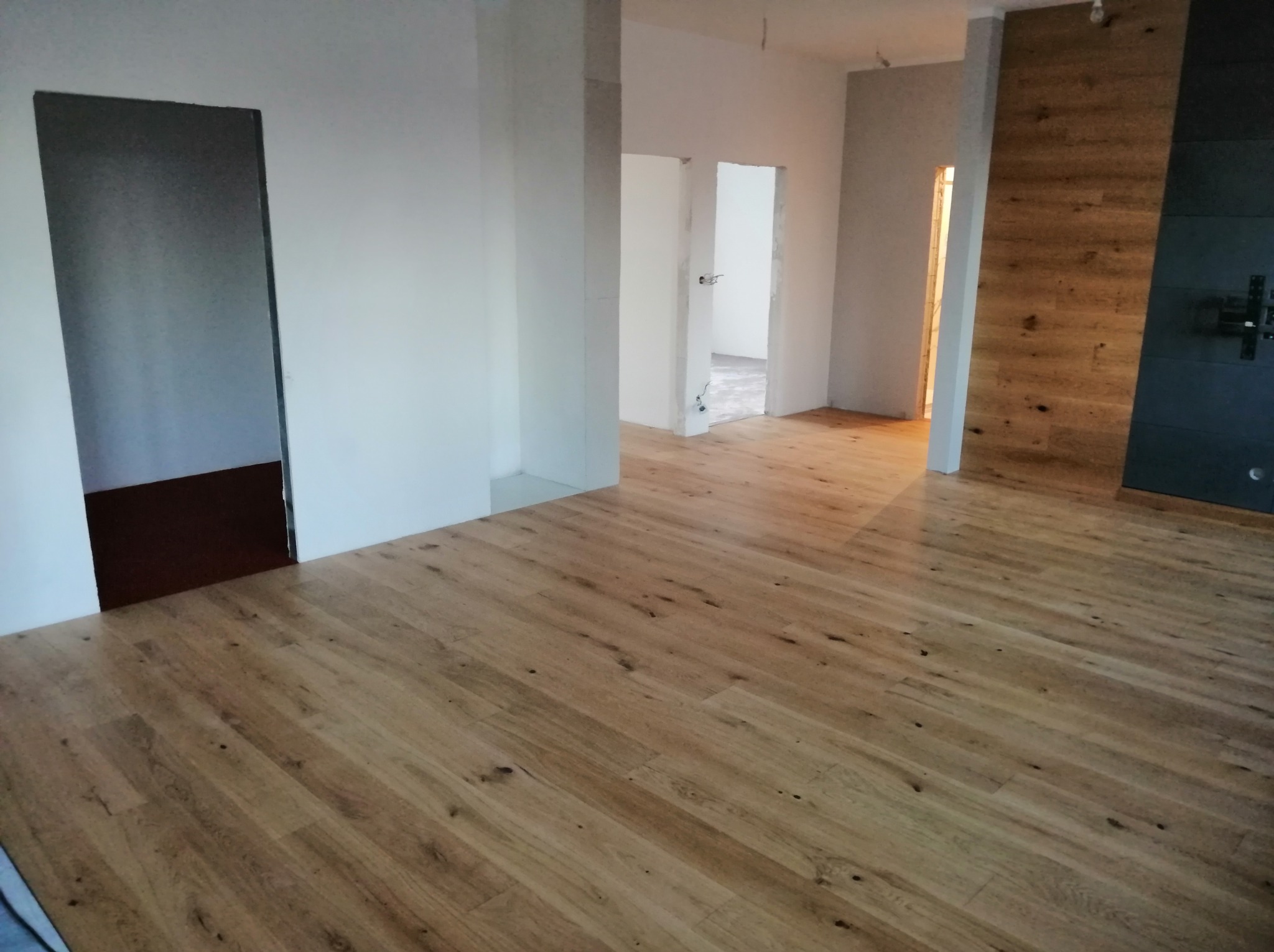 Jasny parkiet ułożony w przestronnym, odświeżonym wnętrzu z białymi ścianami i ciemnymi akcentami, widoczne przejścia do kolejnych pomieszczeń.