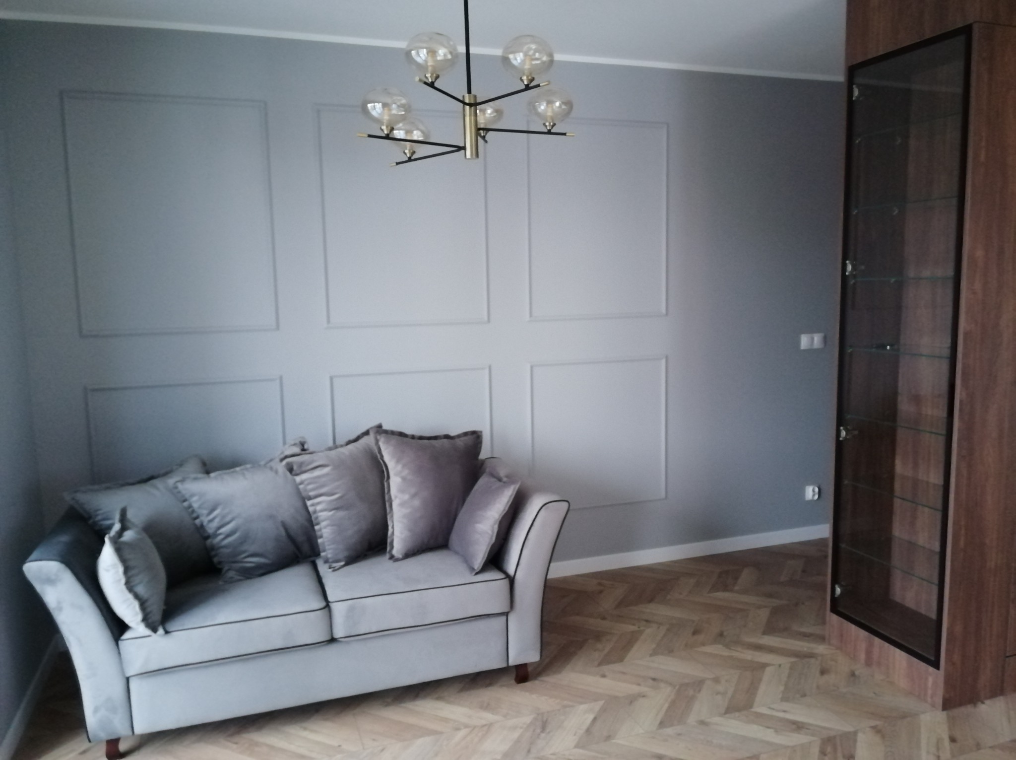 Jasnoszary salon z dekoracyjnymi panelami ściennymi, szarą sofą z poduszkami, nowoczesną lampą sufitową i drewnianą podłogą ułożoną w jodełkę.