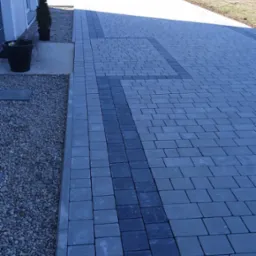 Szara kostka brukowa ułożona w regularny wzór z ciemniejszym pasem wzdłuż podjazdu, obok domu z dekoracyjnymi roślinami w donicach i żwirowym obrzeżem.