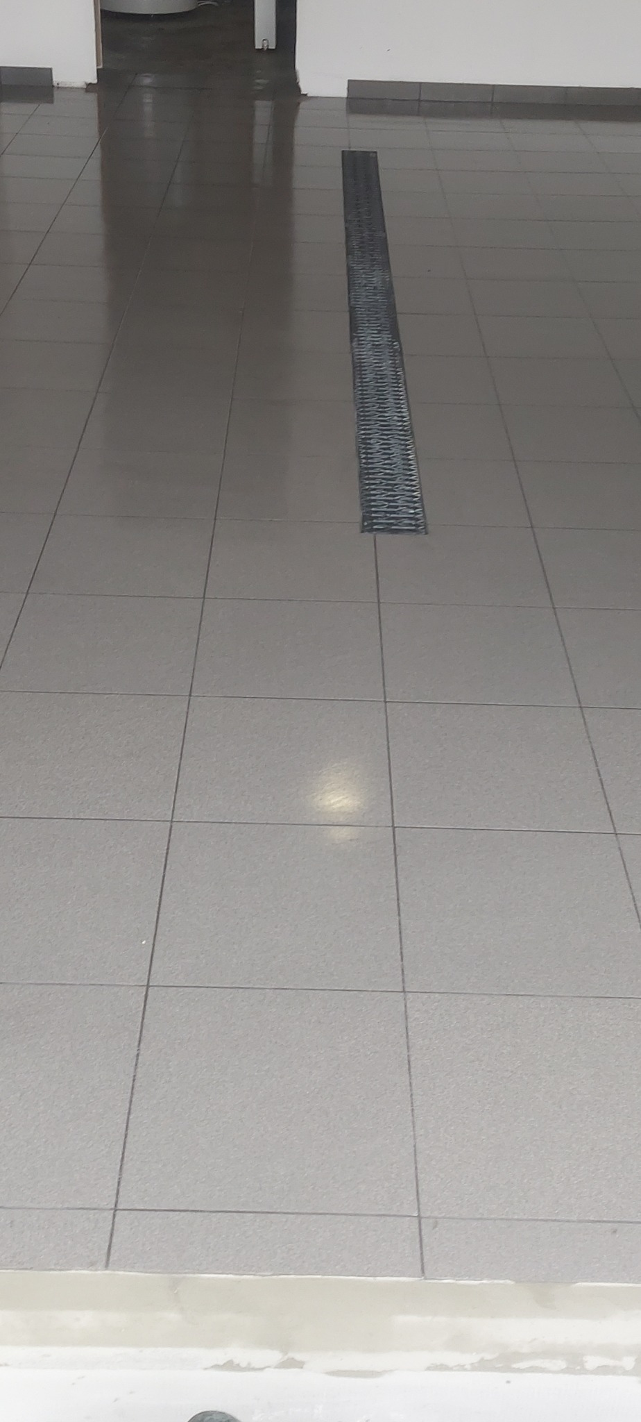 Szare płytki podłogowe z metalowym odpływem liniowym, widoczne fugi i odbicie światła, w tle drzwi.