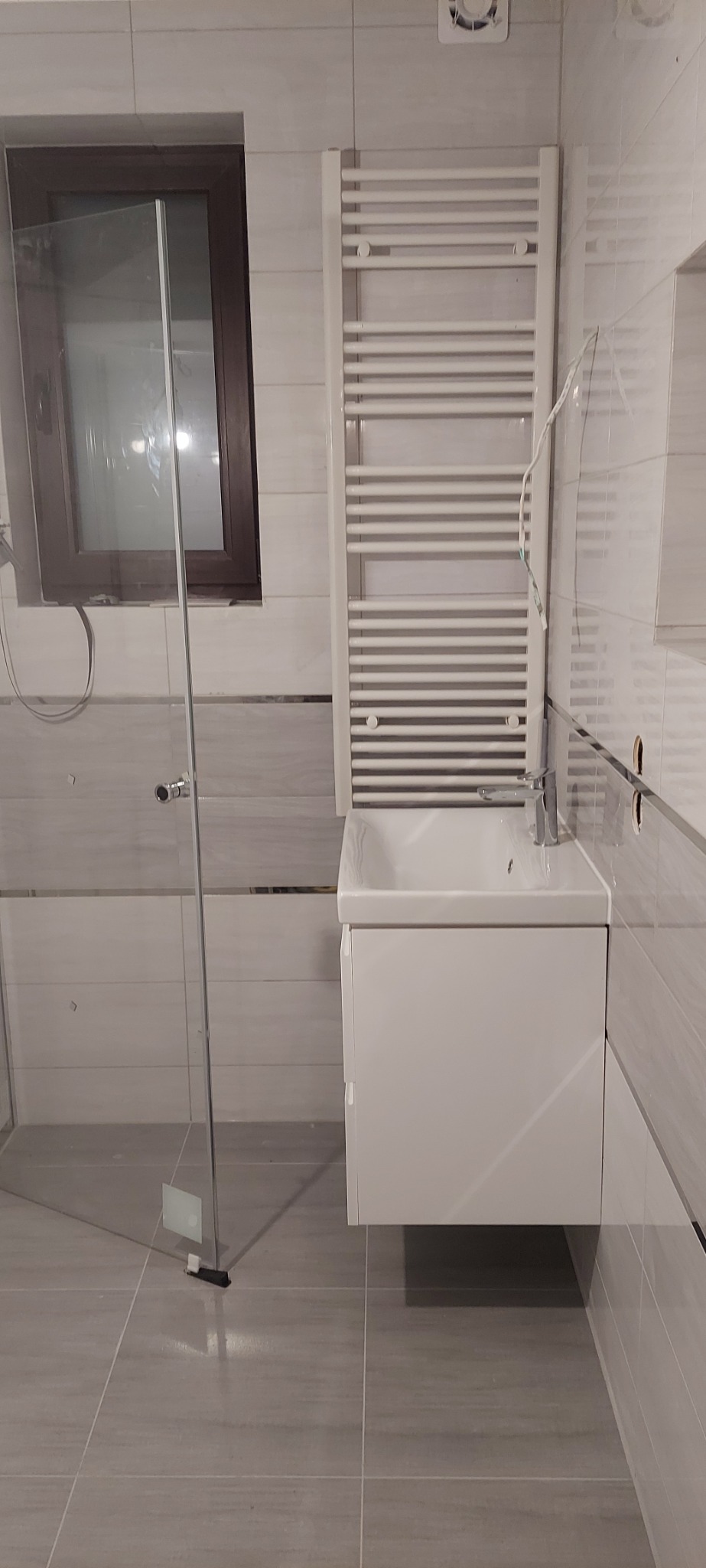 Wykończona łazienka z kabiną prysznicową ze szklanymi drzwiami, umywalką z szafką w kolorze białym i grzejnikiem drabinkowym na tle szarych płytek ceramicznych z delikatnym wzorem.