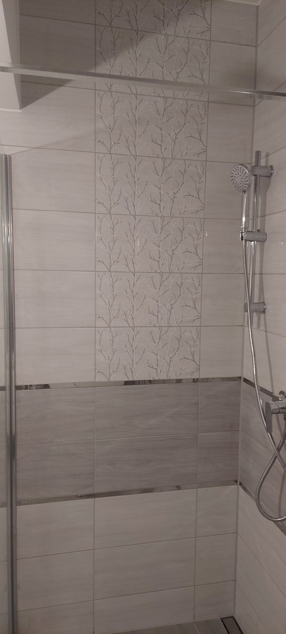 Wykończona kabina prysznicowa w Przybiernowie z beżowymi płytkami, w górnej części dekoracyjna mozaika z motywem drzewa, widoczna słuchawka prysznicowa.