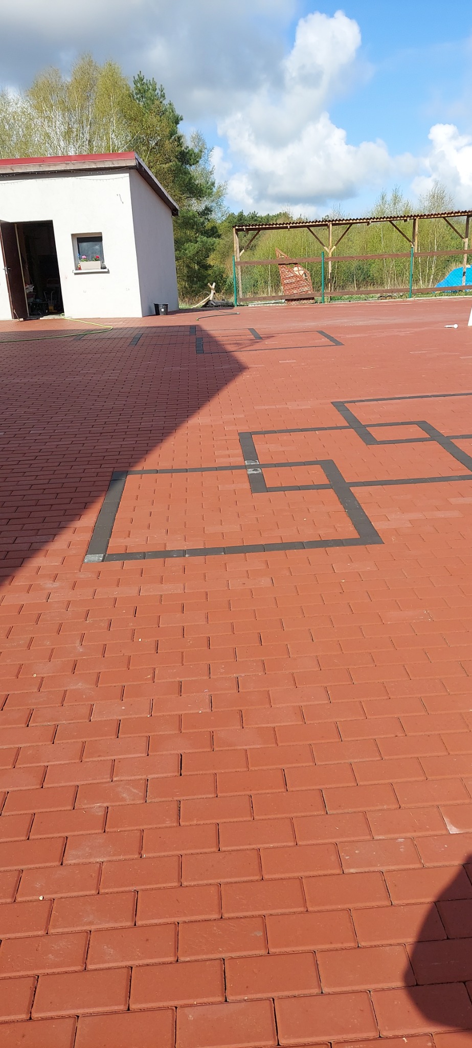 Czerwona kostka brukowa ułożona w geometryczny wzór z ciemnoszarymi obramowaniami, widoczny budynek gospodarczy i zadaszenie w tle.