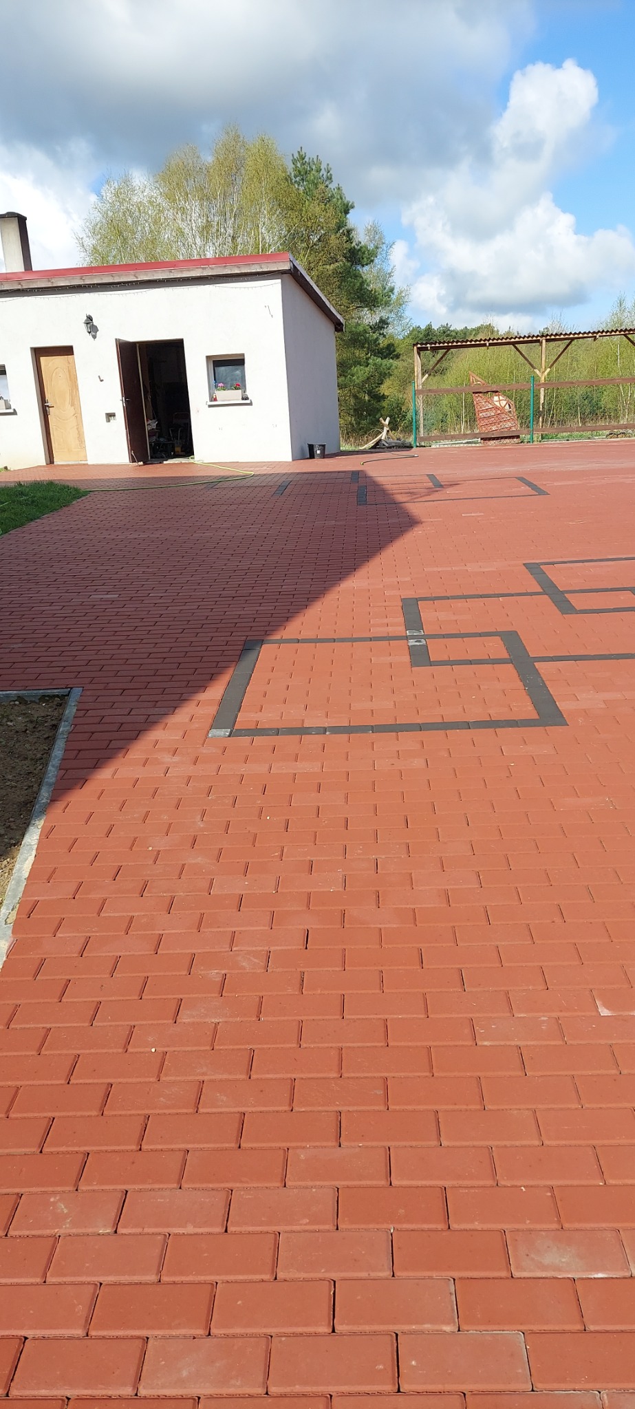 Nowo ułożona, czerwona kostka brukowa z czarnymi obramowaniami tworzącymi geometryczne wzory na podwórku z budynkiem gospodarczym i zadaszoną konstrukcją w tle.