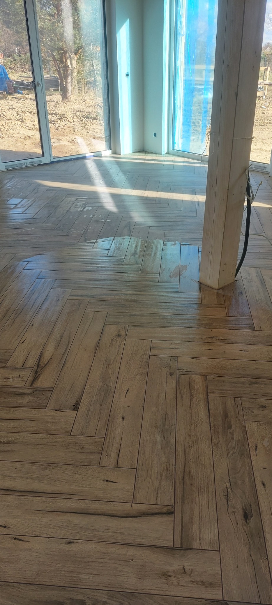 Ułożona w jodełkę podłoga z płytek imitujących drewno, w jasnym wnętrzu z dużymi oknami, częściowo zasłoniętymi folią ochronną, z widocznym drewnianym słupem i okablowaniem.