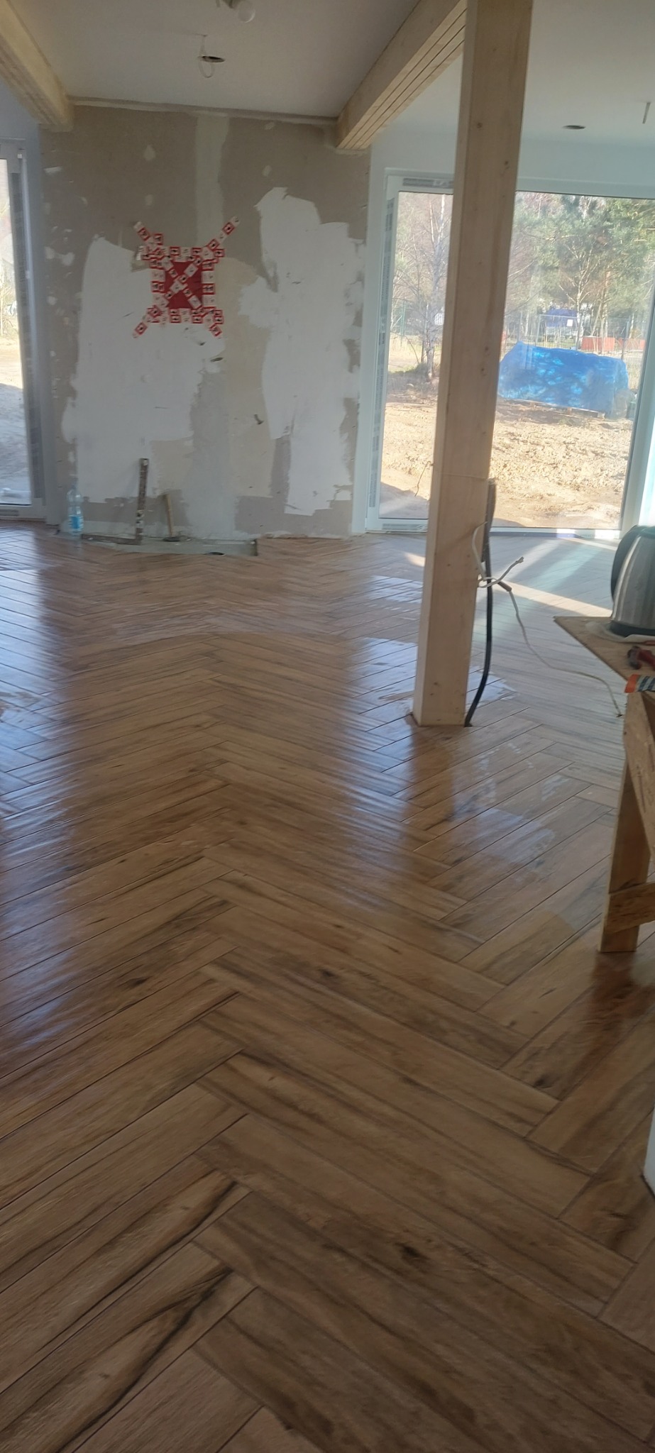 Ułożona w jodełkę, błyszcząca podłoga z paneli drewnopodobnych w trakcie remontu, z widoczną surową ścianą i słupem konstrukcyjnym w tle.