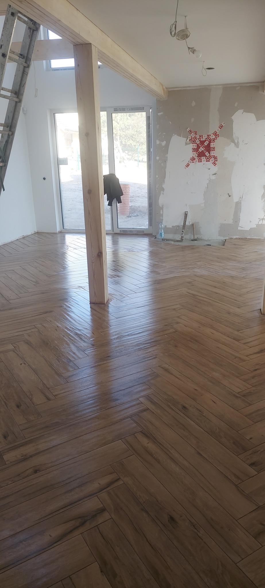 Ułożona podłoga z płytek imitujących drewno w jodełkę, widoczne narzędzia, nieotynkowana ściana z zaznaczonymi punktami, drewniany słup podtrzymujący strop, drabina i okno balkonowe.