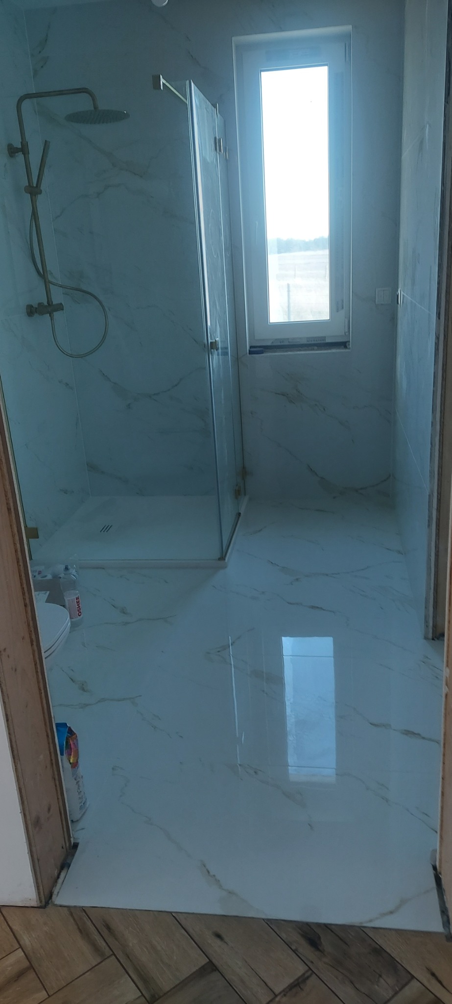 Nowoczesna łazienka z kabiną prysznicową typu walk-in, wykończona białymi płytkami imitującymi marmur, z złotą armaturą i oknem w tle.