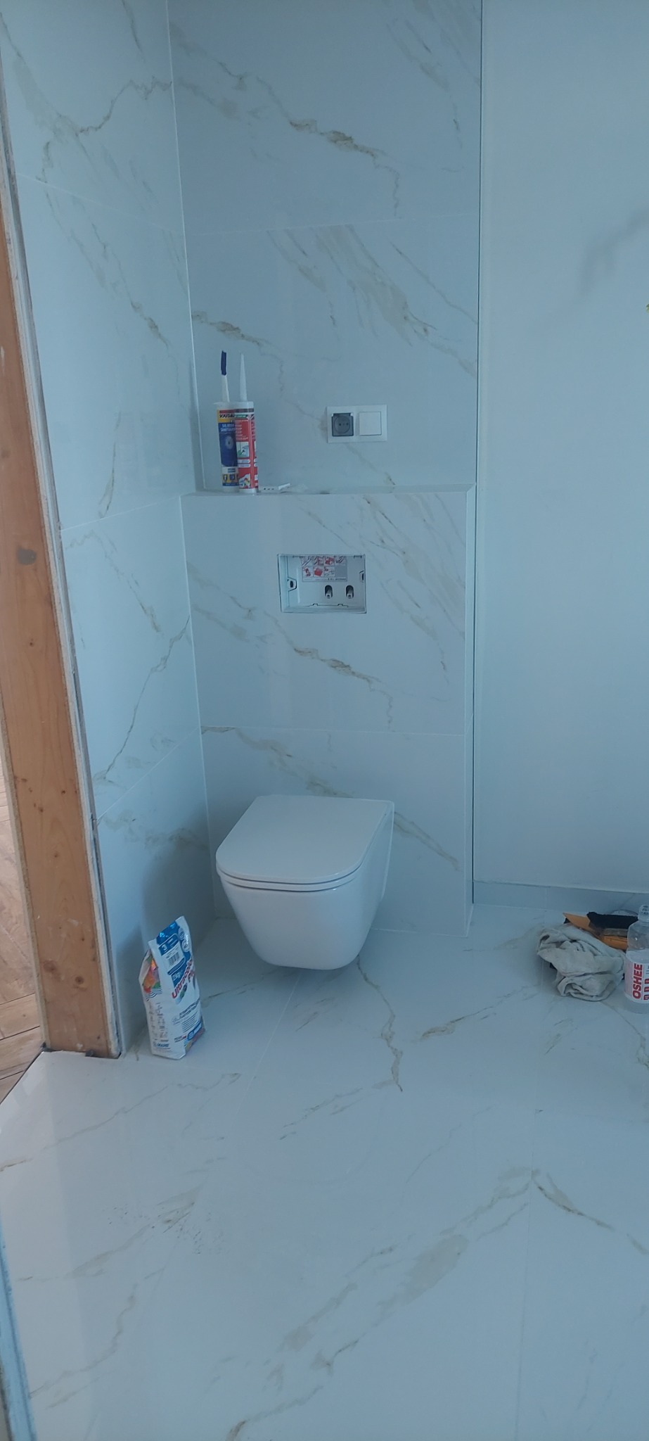 Świeżo wykończona łazienka z podwieszaną toaletą na tle płytek imitujących marmur, widoczne akcesoria montażowe i zaprawa w worku.