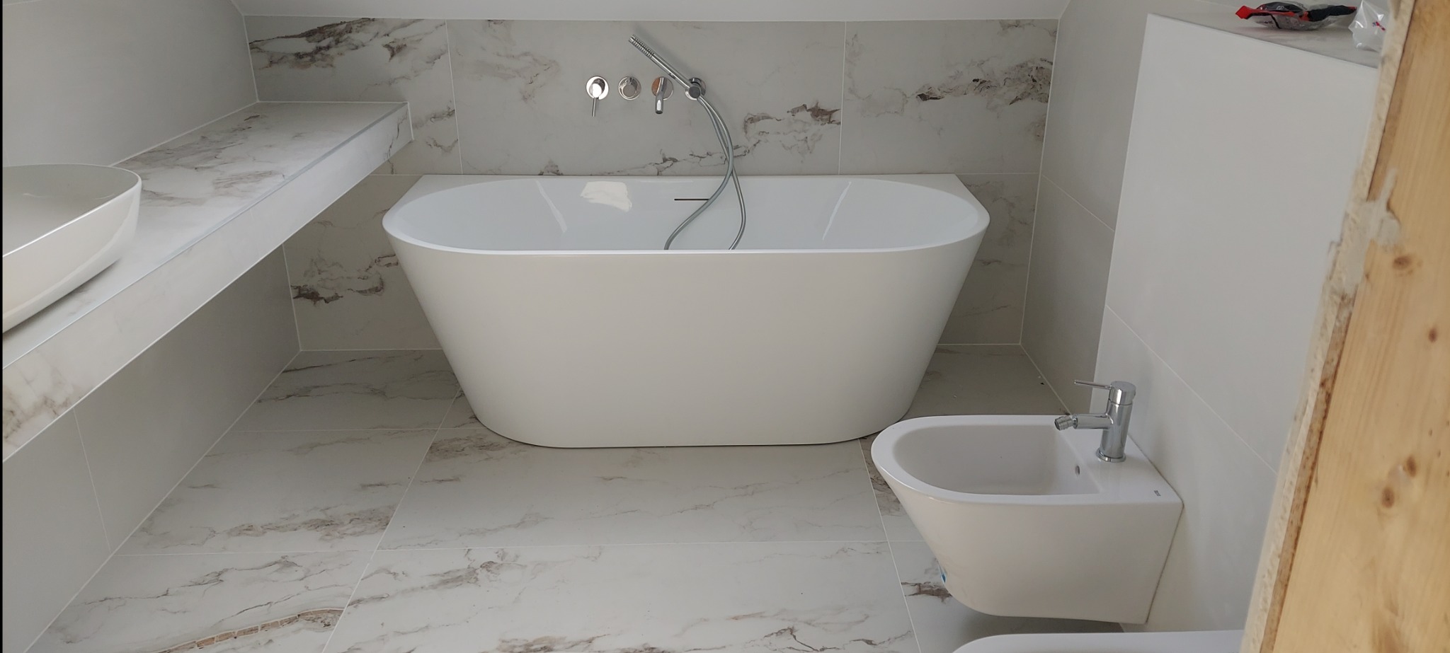 Nowoczesna łazienka z wolnostojącą wanną, bidetem i umywalką nablatową na blacie z płytek imitujących marmur, wykończona płytkami w podobnym stylu, widok na chromowaną armaturę i fragment...