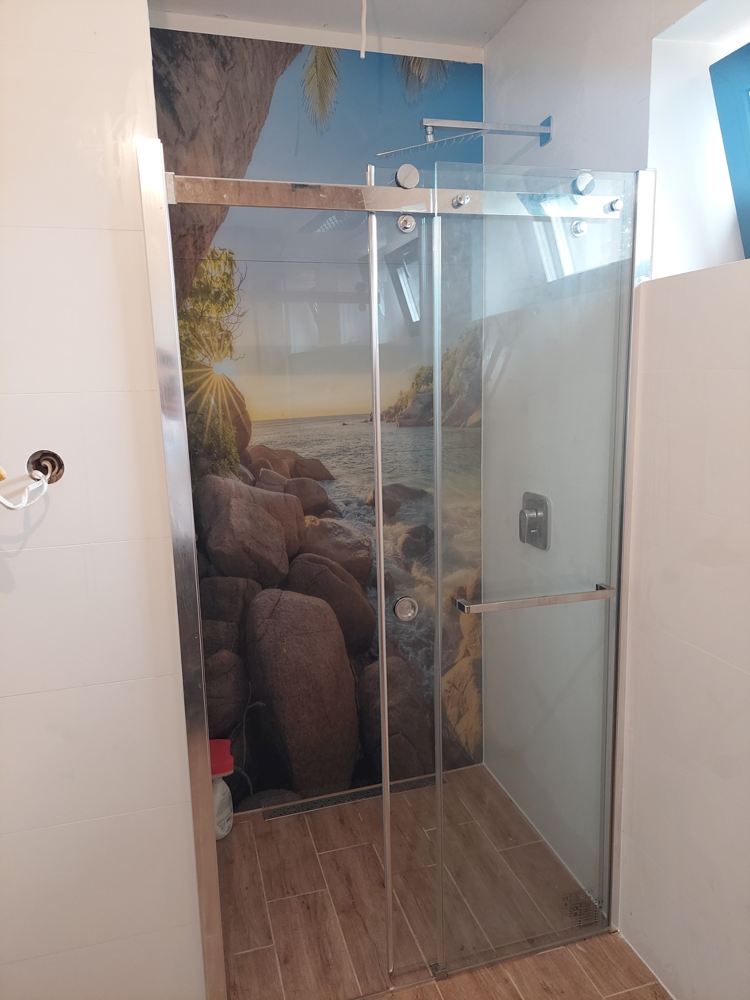 Szklana kabina prysznicowa z przesuwnymi drzwiami, na których znajduje się grafika przedstawiająca plażę z kamieniami i zachodzące słońce, zamontowana w łazience z białymi płytkami i drewnopodobną...