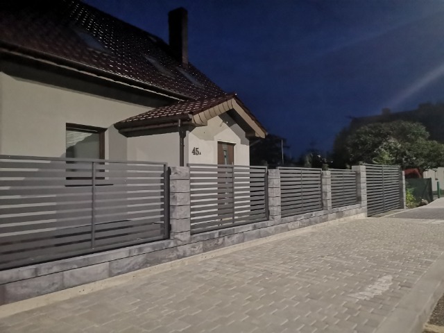 Nowoczesne ogrodzenie panelowe w kolorze szarości z betonowymi słupkami i podmurówką, oświetlone nocnym światłem, widoczne z chodnika przed domem z numerem 45.