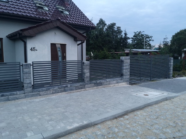 Nowoczesne, poziome ogrodzenie z szarych paneli i bloków betonowych przed domem z numerem 45 i brukowanym chodnikiem.