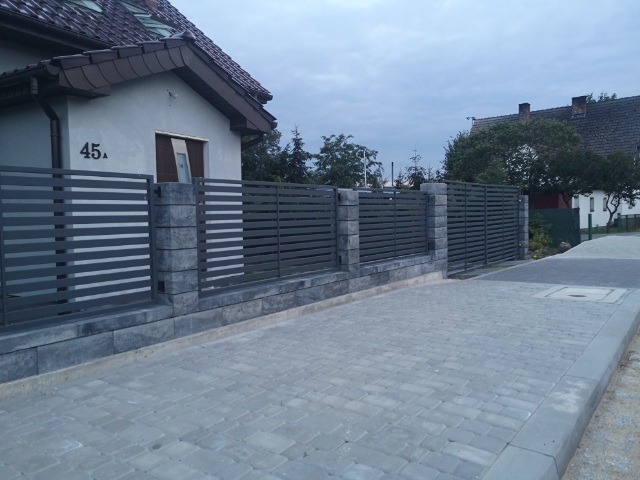 Nowoczesne ogrodzenie panelowe w kolorze grafitowym z podmurówką i słupkami z szarej kostki, przylegające do domu z numerem 45a i brukowanym podjazdem.