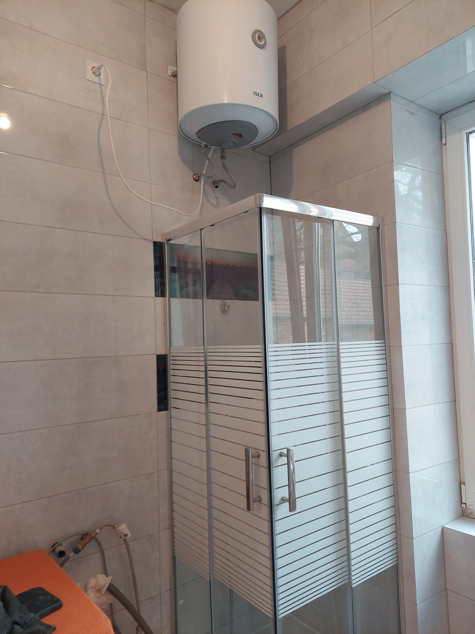 Zamontowana kabina prysznicowa ze szklanymi drzwiami z poziomymi paskami, podgrzewacz wody marki ISEA na ścianie z jasnych płytek.