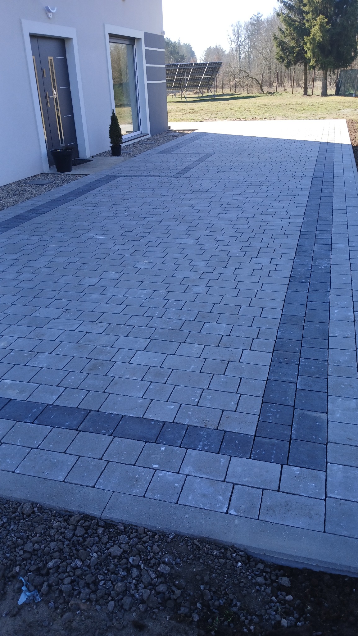 Ułożona szara kostka brukowa z ciemniejszym obramowaniem przed nowoczesnym domem z panelami słonecznymi w tle.