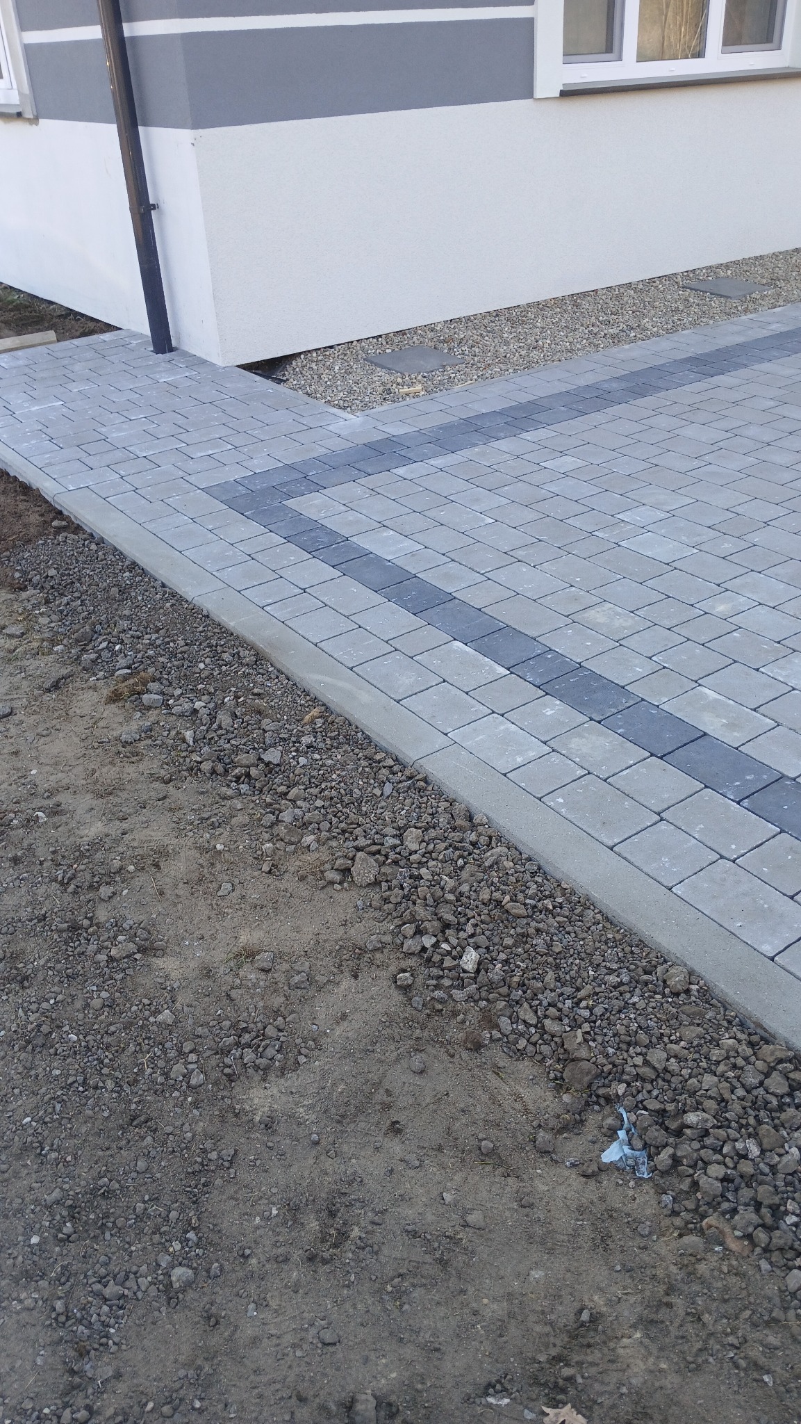 Ułożona szara kostka brukowa z ciemniejszym pasem, obrzeżem i podsypką żwirową przy ścianie budynku.