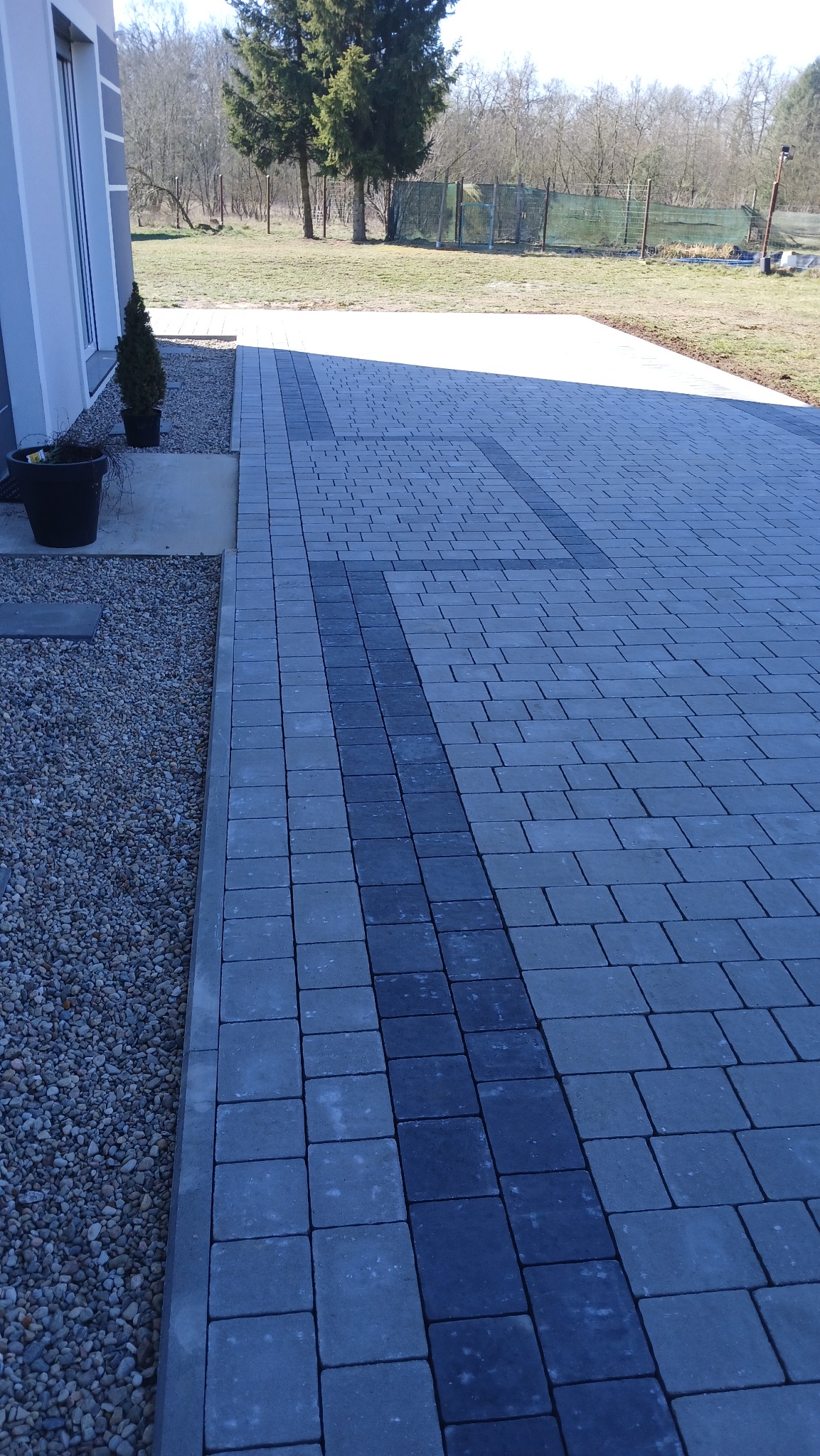 Szara kostka brukowa ułożona w regularny wzór z ciemniejszym pasem wzdłuż podjazdu, obok domu z dekoracyjnymi roślinami w donicach i żwirowym obrzeżem.