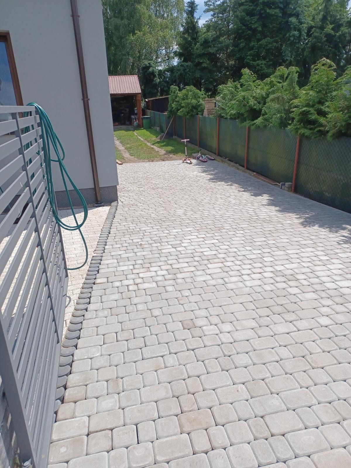 Ułożona szara kostka brukowa na podjeździe, widoczny fragment szarego ogrodzenia panelowego, zielony wąż ogrodowy zwinięty przy ścianie domu, w tle zieleń i budynek gospodarczy z brązowym dachem.
